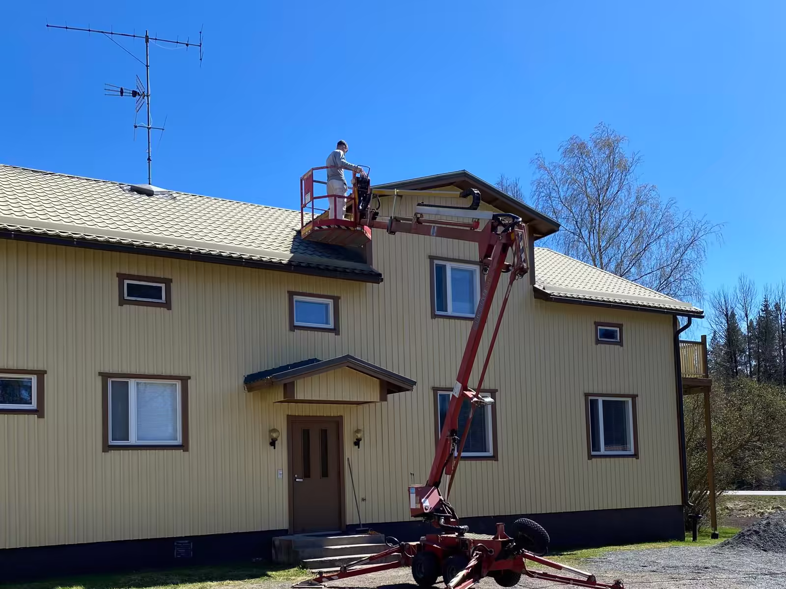 Maalari nostokorissa maalaamassa keltaisen puutalon julkisivua räystään korkeudelta
