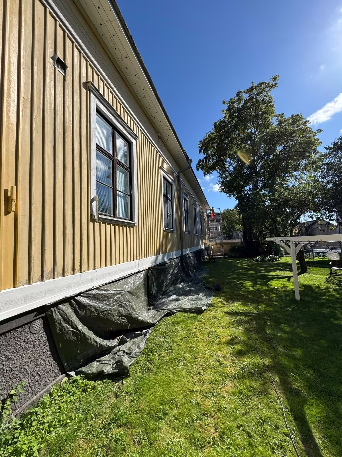 Pitkän puutalon sivuseinä ennen maalausta, kulunut keltainen lautaverhoilu ja suojapeitteet sokkelin edessä